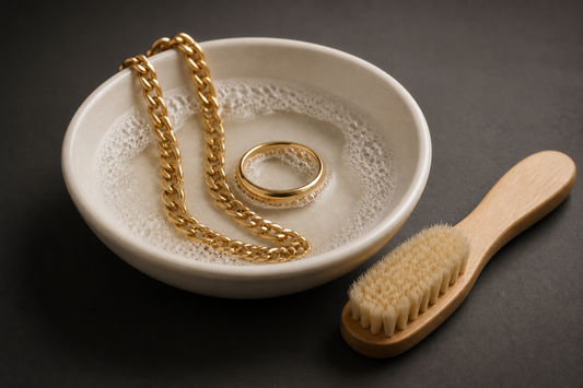 A gold chain and gold ring in a white dish of soapy water beside a soft cleaning brush
